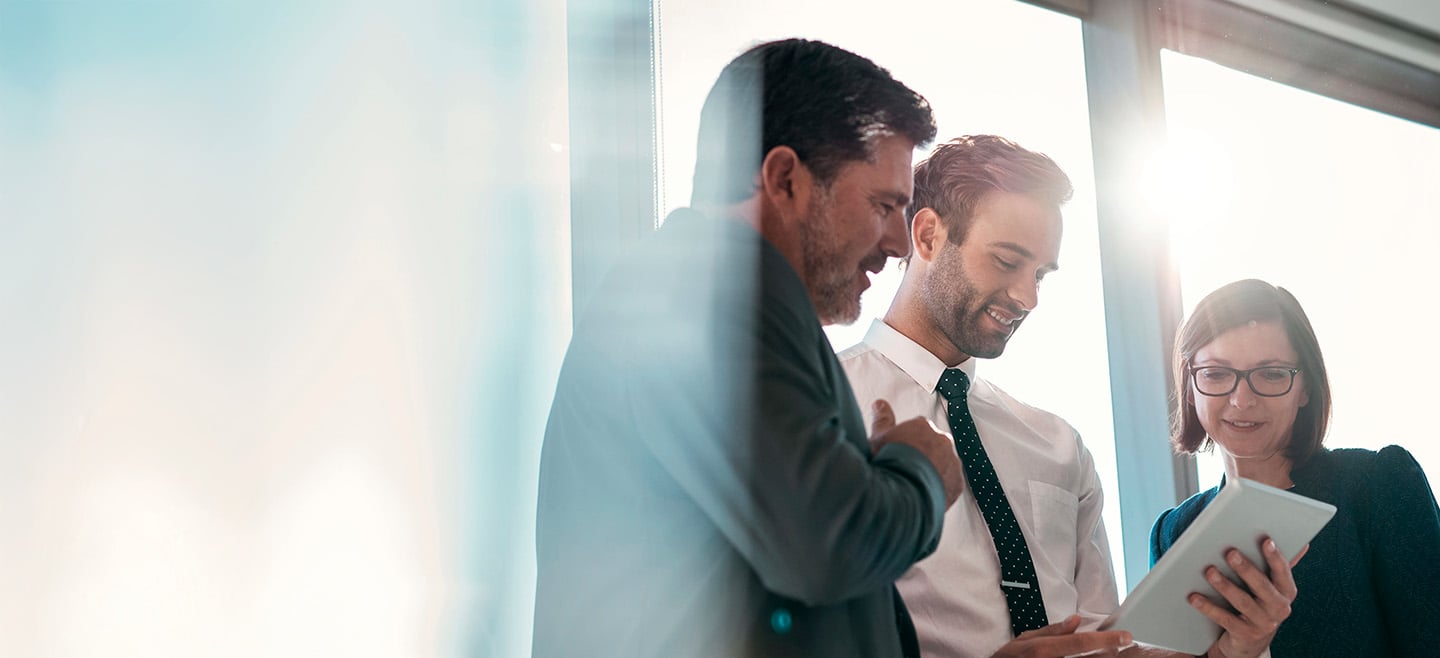 Three business professionals, two men in suits and a woman in a blazer, standing in front of large office windows with sunlight streaming in, looking at and discussing something on a digital tablet with smiles and engagement.
