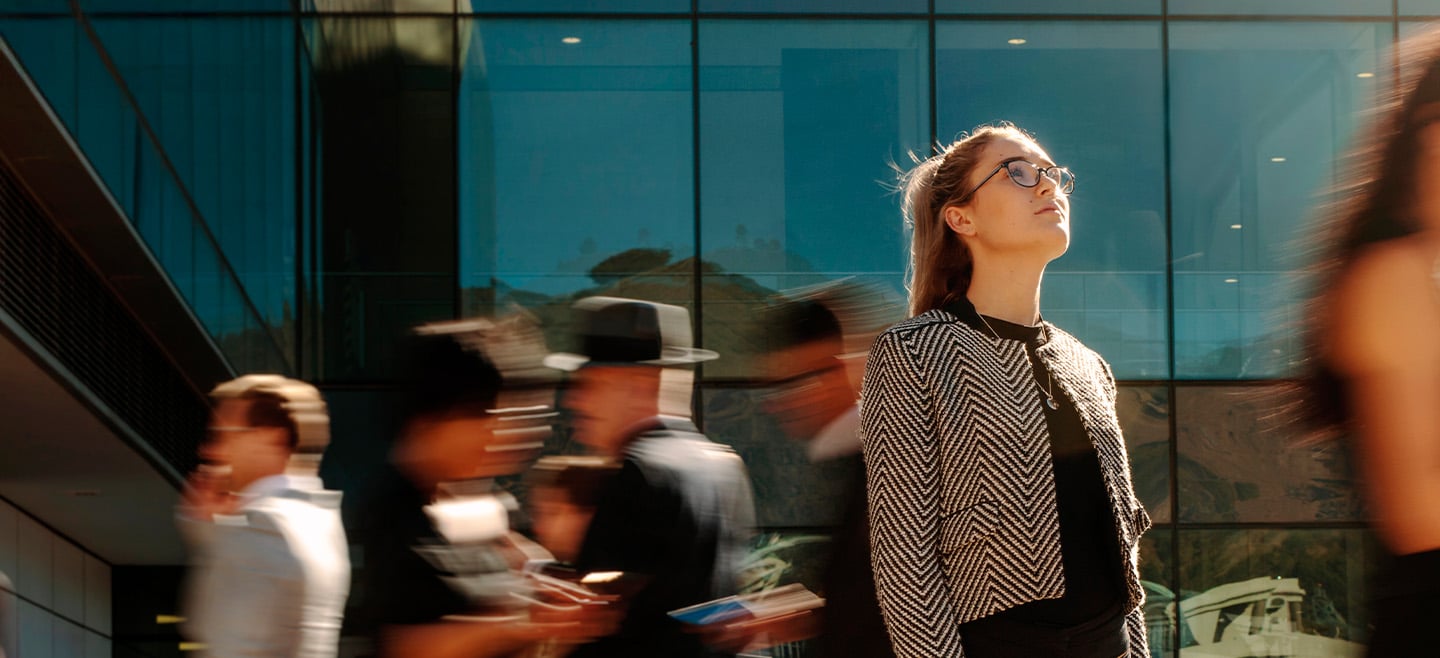 Young woman wearing glasses and a patterned jacket looks upward with a thoughtful expression, standing outdoors against a modern glass building, while blurred figures move past her in the background.