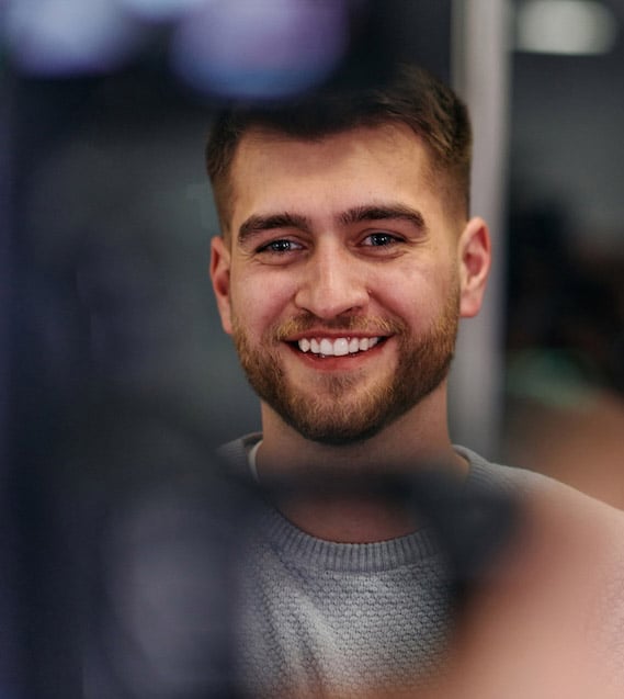 Smiling young man with a neatly trimmed beard wearing a light gray sweater, looking slightly past the camera in a softly lit indoor setting.