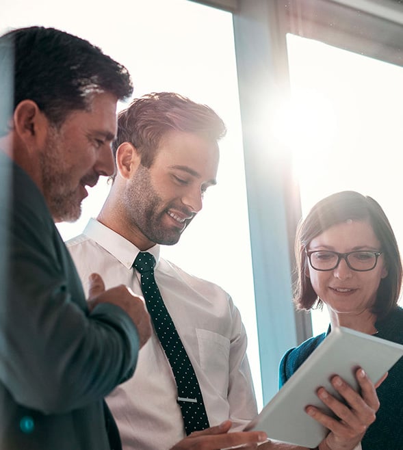 Three business professionals, two men in suits and a woman in a blazer, standing in front of large office windows with sunlight streaming in, looking at and discussing something on a digital tablet with smiles and engagement.